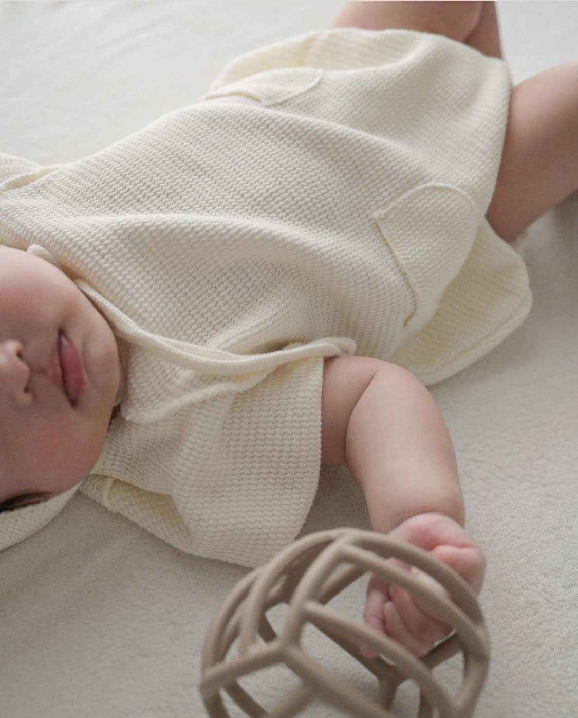 Baby trägt einen cremefarbenen Strick-Hoodie-Strampler von Little Foxx, spielt mit einer Holzrassel – hochwertige Babykleidung für Komfort und Stil.