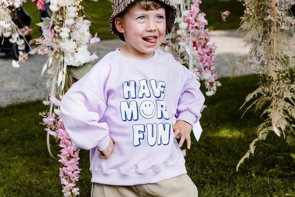 Junge trägt lila Sweatshirt von Little Foxx mit ‚Have More Fun‘-Print und Bucket Hat, umgeben von Blumenarrangements im Freien.