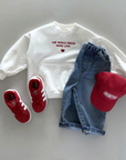 Weißes Sweatshirt mit rotem Schriftzug, kombiniert mit einer roten Cap, Jeansrock und roten Sneakern.
