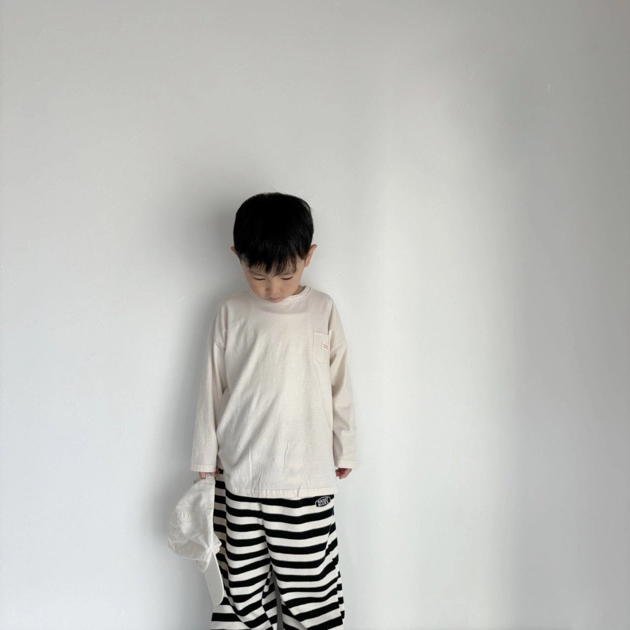 Kind trägt das cremefarbene Longsleeve-Tee mit einer schwarz-weiß gestreiften Hose.