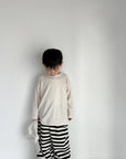 Kind trägt das cremefarbene Longsleeve-Tee mit einer schwarz-weiß gestreiften Hose.