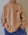 Kind trägt das beige „Frosted Flakes“ Sweatshirt – kleiner roter „Kellogg’s“ Print auf der Brust sichtbar.