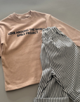 Beiges Longsleeve mit Schriftzug „LOVE AROUND THE WORLD“ zusammen mit einer gestreiften Hose.