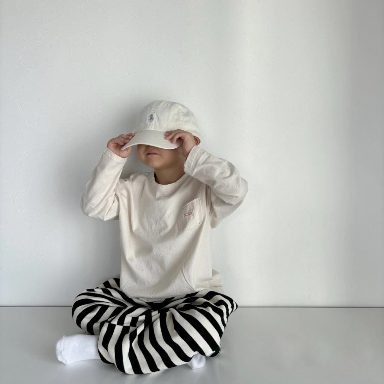 Kind im cremefarbenen Longsleeve-Tee mit gestreifter Hose und Cap – lässiger Look.