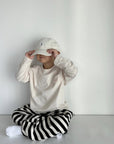 Kind im cremefarbenen Longsleeve-Tee mit gestreifter Hose und Cap – lässiger Look.