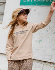 Kind in beigem Longsleeve mit „LOVE AROUND THE WORLD“ Schriftzug – Bewegungsszene mit lockerer Passform.