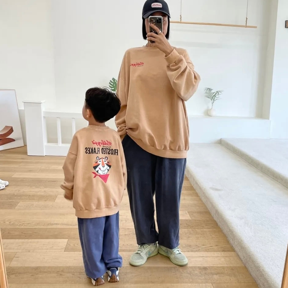 Spiegelbild von Kind und Erwachsenem im beigen „Frosted Flakes“ Sweatshirt – „Kellogg’s“ Schriftzug vorne, großer Print mit Tony der Tiger auf dem Rücken.