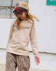 Kind trägt das beige Longsleeve mit schwarzem Schriftzug – lässiger Look mit Leo Hose.