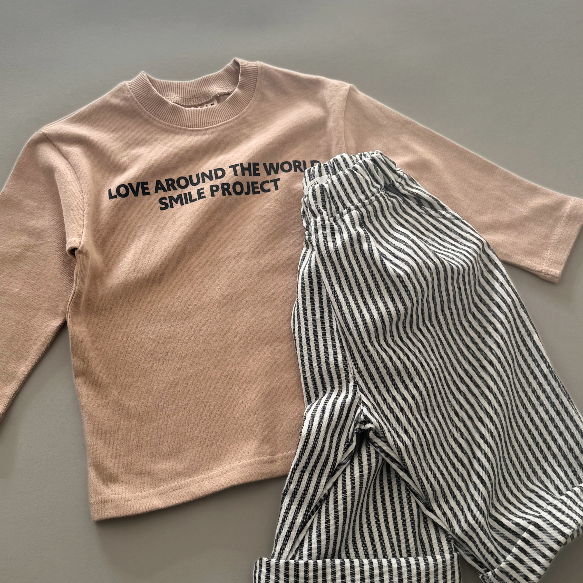 Beiges Longsleeve mit Schriftzug „LOVE AROUND THE WORLD“ zusammen mit einer gestreiften Hose.