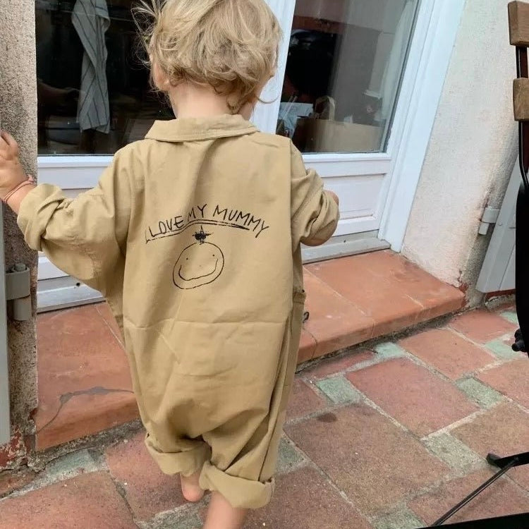 Kleines Kind in beigem Statement-Overall aus Baumwolle mit „I Love My Mummy“-Print auf dem Rücken, barfuß auf einer Terrasse – exklusiv bei Little Foxx Concept Store.