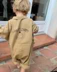 Kleines Kind in beigem Statement-Overall aus Baumwolle mit „I Love My Mummy“-Print auf dem Rücken, barfuß auf einer Terrasse – exklusiv bei Little Foxx Concept Store.