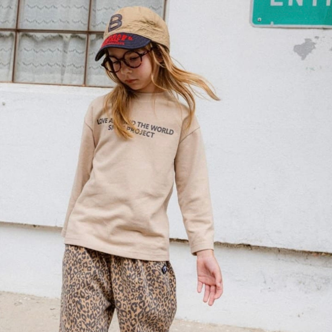 Kind trägt das beige Longsleeve mit schwarzem Schriftzug – lässiger Look mit Leo Hose.