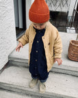 Kleines Kind mit orangefarbener Mütze und beiger Steppjacke trägt einen dunkelblauen Denim-Overall und beige Schnürschuhe, steht auf Treppen vor einem Hauseingang mit geflochtenen Körben.