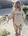 Kind mit schulterlangen Haaren läuft auf einem sandigen Pfad durch eine sonnige Landschaft in Sylt, trägt einen beigen Statement-Overall mit „I Love My Mummy“-Print und braune Schuhe.