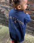 Kind in dunkelblauem Denim-Overall mit „I Love My Mummy“-Print auf dem Rücken steht vor einer alten Backsteinmauer und schaut zur Seite, entspannter Streetstyle-Look.