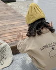 Mädchen in beigem Statement-Overall aus Baumwolle mit „I Love My Mummy“-Print auf dem Rücken, gelber Mütze und Stofftasche, kniend auf einer Holzterrasse – exklusiv bei Little Foxx Concept Store.